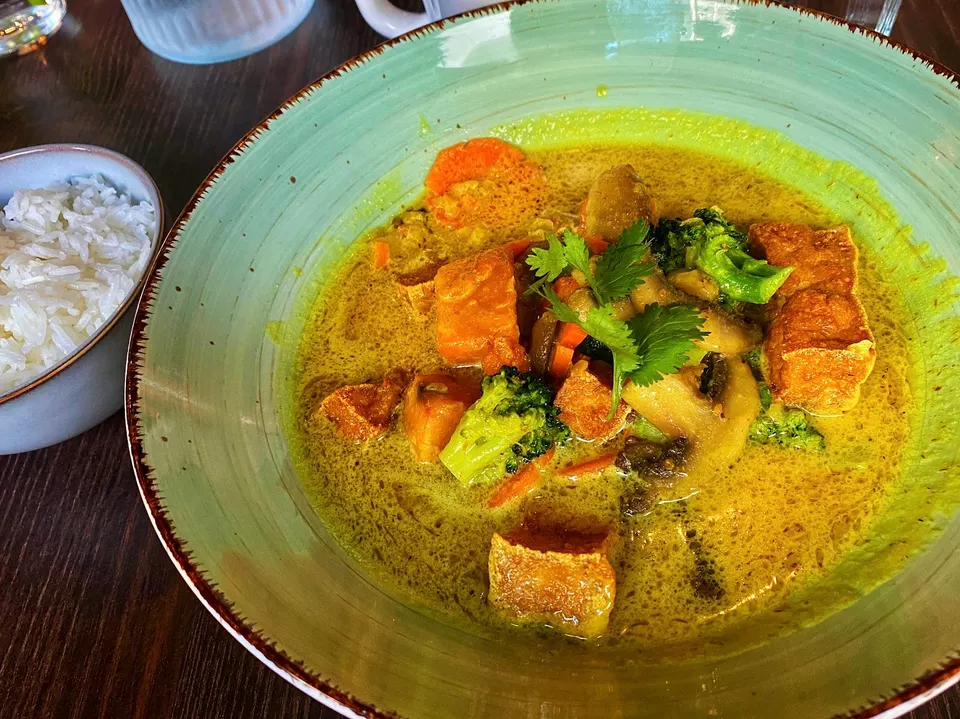 A bowl of yellow curry with tofu, broccoli, carrots, and mushrooms, garnished with cilantro, served alongside a small bowl of white rice on a dark wooden table.