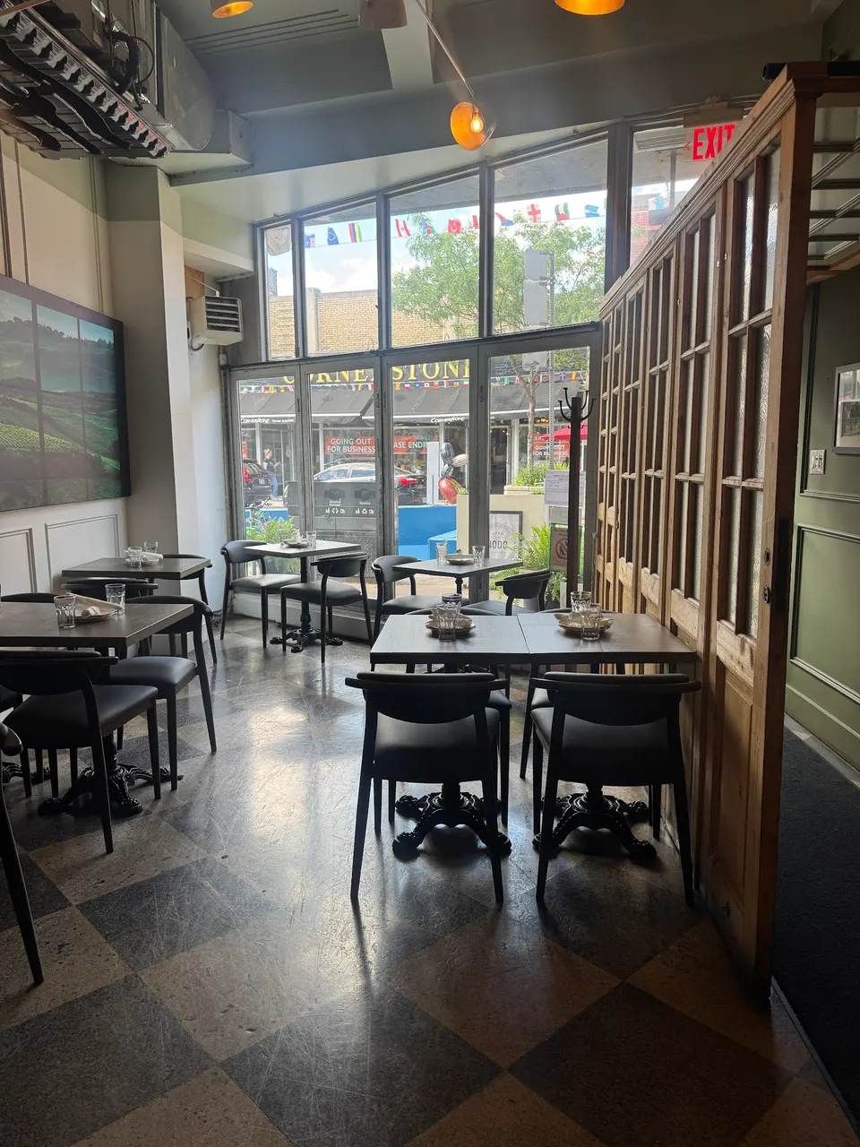 An indoor dining area of a restaurant featuring dark wooden tables and chairs, a checkered floor, large windows looking out onto a street, and a wooden partition wall.