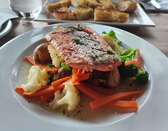 A grilled salmon fillet served over a bed of roasted potatoes, carrots, cauliflower, and broccoli in a light sauce on a white plate, with garlic bread in the background.