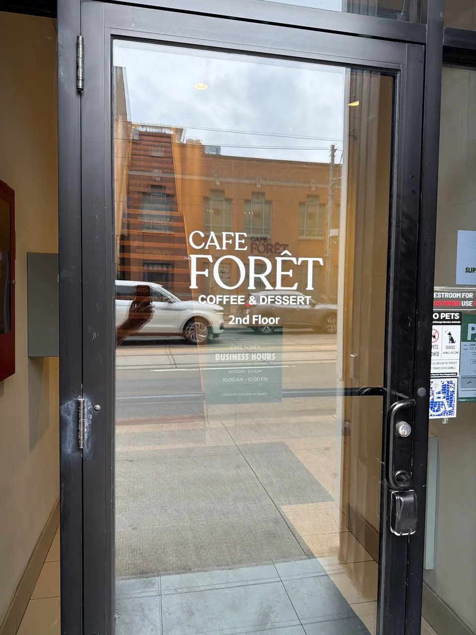 A glass entrance door with white lettering that reads 'CAFE FORÊT COFFEE & DESSERT 2nd Floor' and lists business hours.