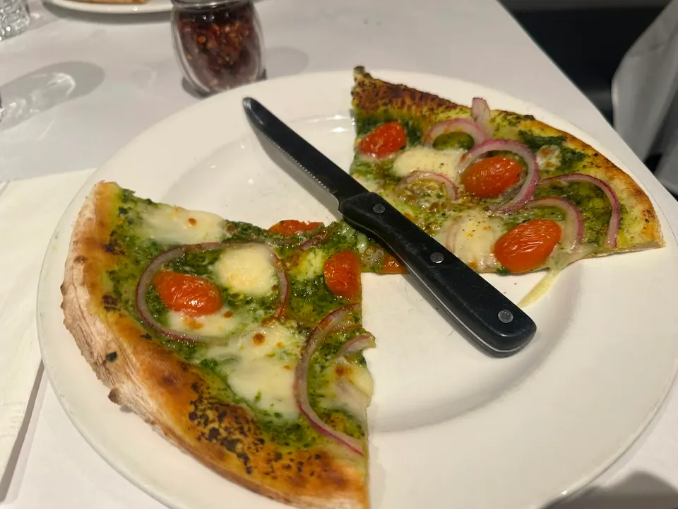 Two slices of pesto pizza topped with melted mozzarella, cherry tomatoes, and red onion rings on a white plate with a black-handled knife.