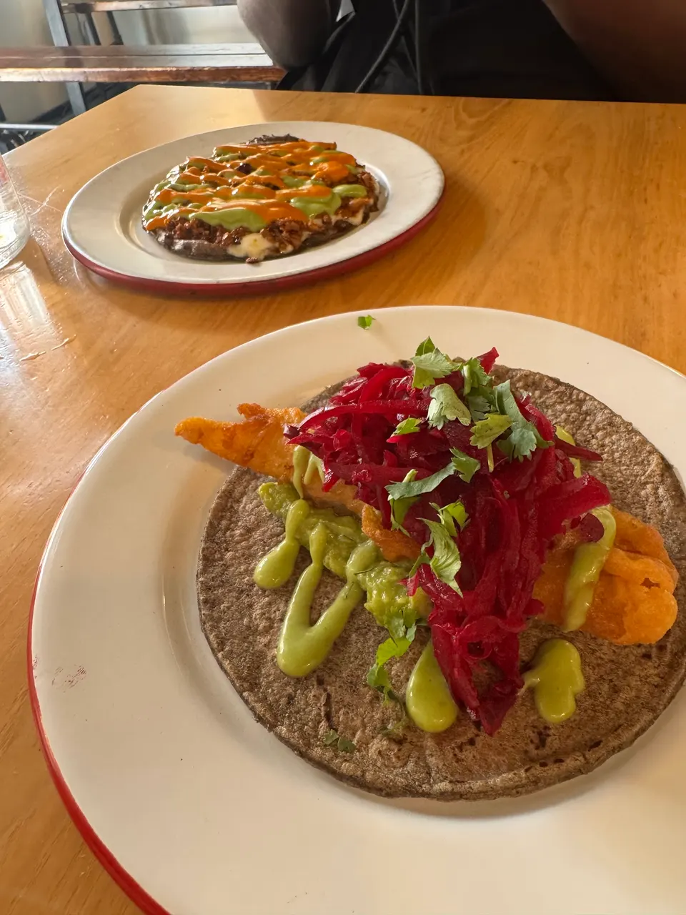 A close-up of a fish taco on a dark corn tortilla topped with green sauce, pickled red cabbage, and cilantro, with another flatbread dish in the background on a wooden table.
