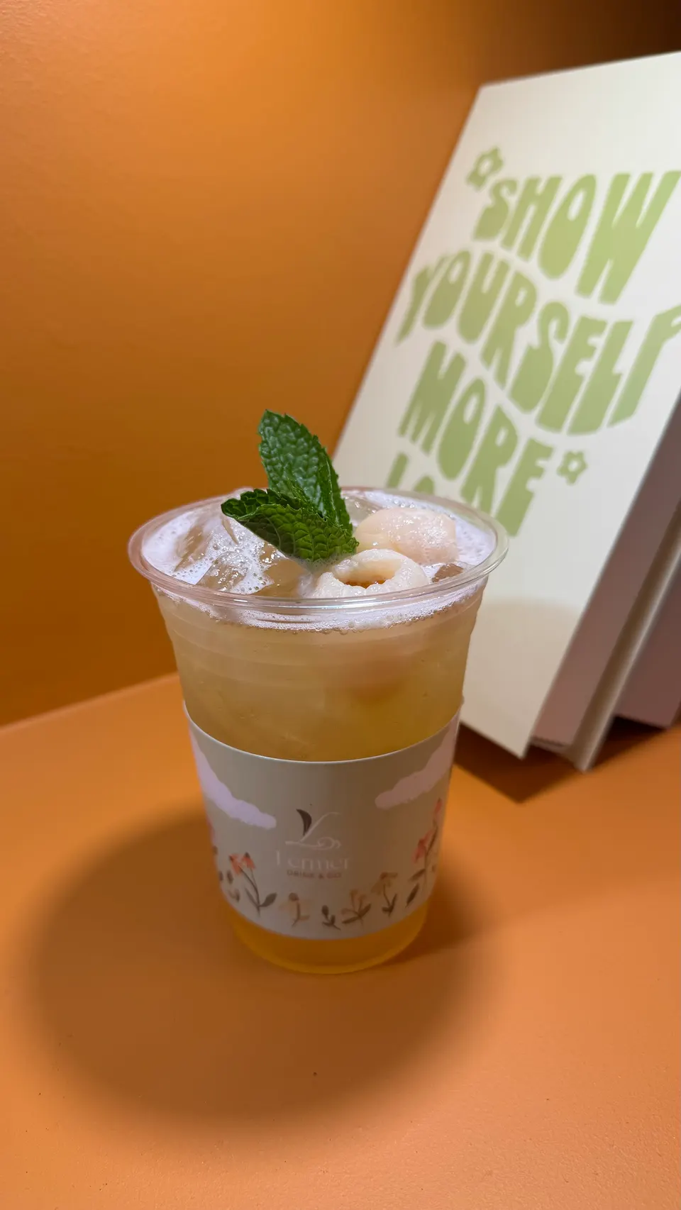 An iced lychee tea drink in a clear plastic cup with a decorative sleeve, garnished with fresh mint leaves and whole lychees, set against an orange background next to a book.