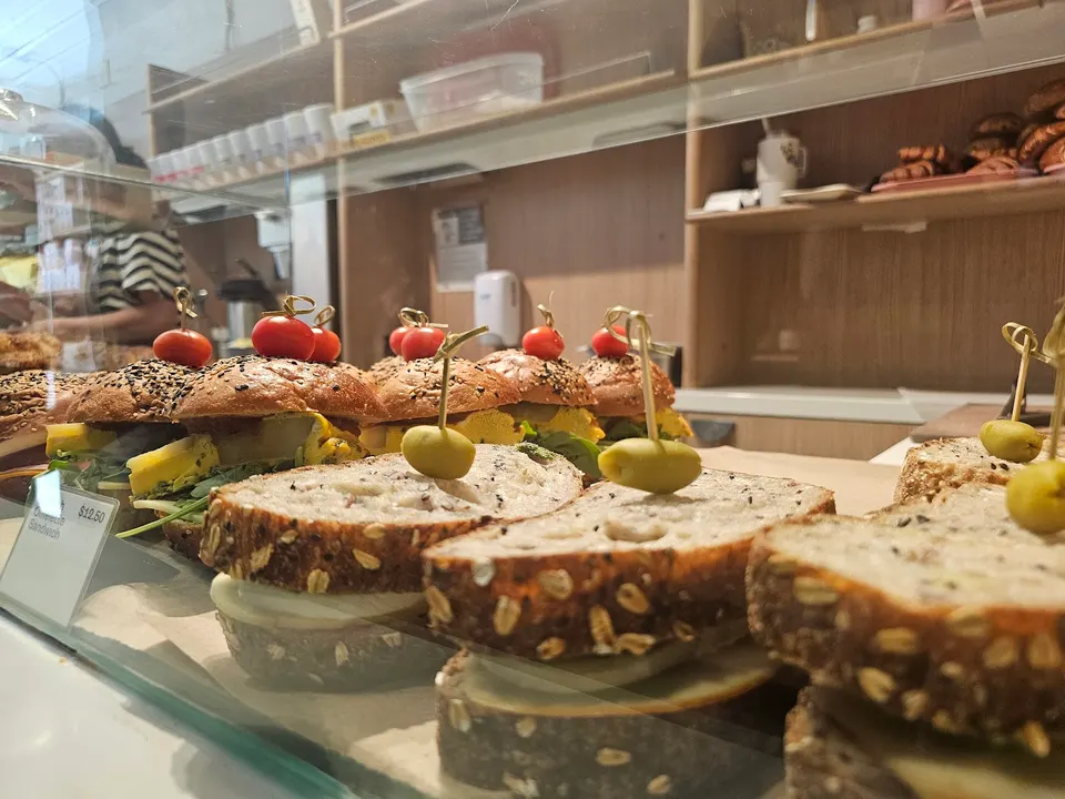 A close-up shot of freshly prepared sandwiches on display behind a glass counter at Dear Grain bakery, featuring seeded bread, olives, and cherry tomatoes on toothpicks.