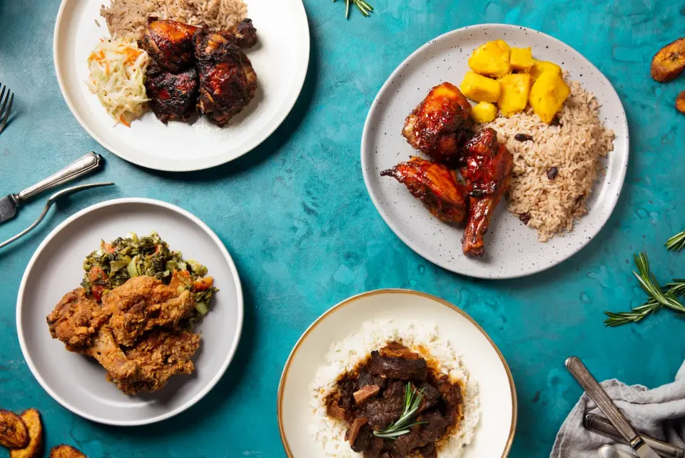 Four plates of Caribbean food are arranged on a blue-green table. One plate has jerk chicken, rice and peas, and coleslaw. Another has jerk chicken, rice and peas, and plantains. A third plate features fried chicken and greens. The fourth plate contains stewed meat over white rice. Utensils and rose
