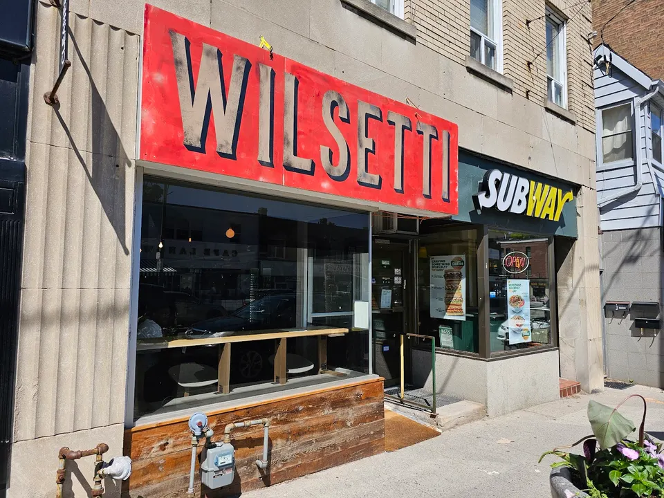 Street view of the Wilsetti storefront with a large red sign featuring bold black lettering, located next to a Subway restaurant on a sunny day.