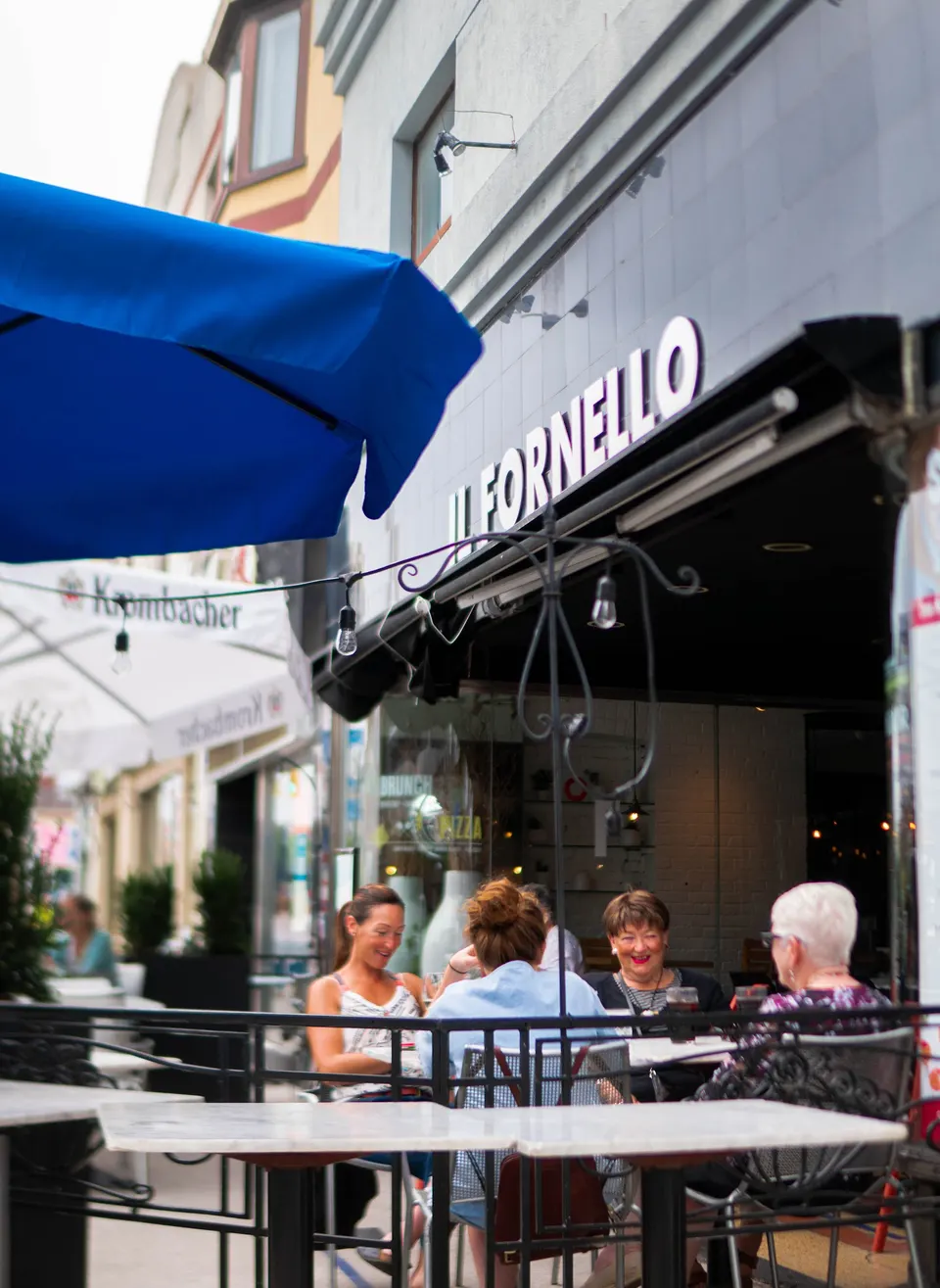 Four women sit at an outdoor patio table under a black awning with the sign 'IL FORNELLO' visible above them, next to a blue umbrella.