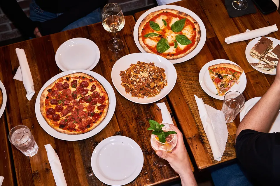 An overhead view of a wooden restaurant table filled with two whole pizzas, a plate of pasta, a slice of pizza, desserts, wine, and water glasses.
