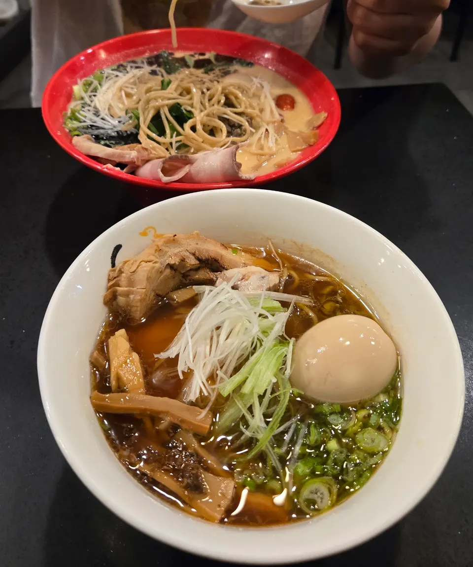 Two bowls of ramen on a black table at Konjiki Ramen, featuring a foreground bowl with dark broth, a soft-boiled egg, bamboo shoots, and green onions, and a background red bowl with creamy broth and noodles.