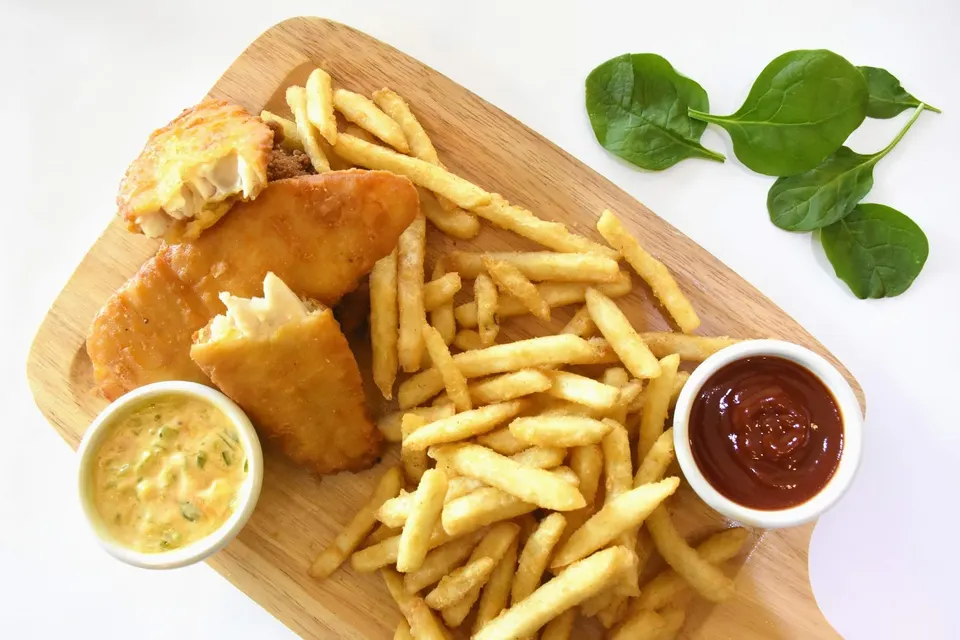A wooden serving board loaded with golden-brown battered fish fillets, a large portion of french fries, a small bowl of tartar sauce, and a small bowl of ketchup, with fresh spinach leaves on the side.