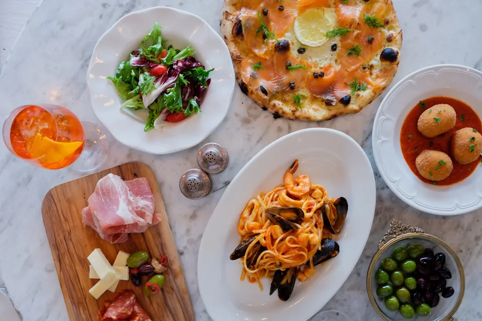 An overhead view of an Italian feast on a marble table, featuring seafood pasta, a smoked salmon pizza, a fresh green salad, arancini in tomato sauce, a charcuterie board, olives, and an orange cocktail.
