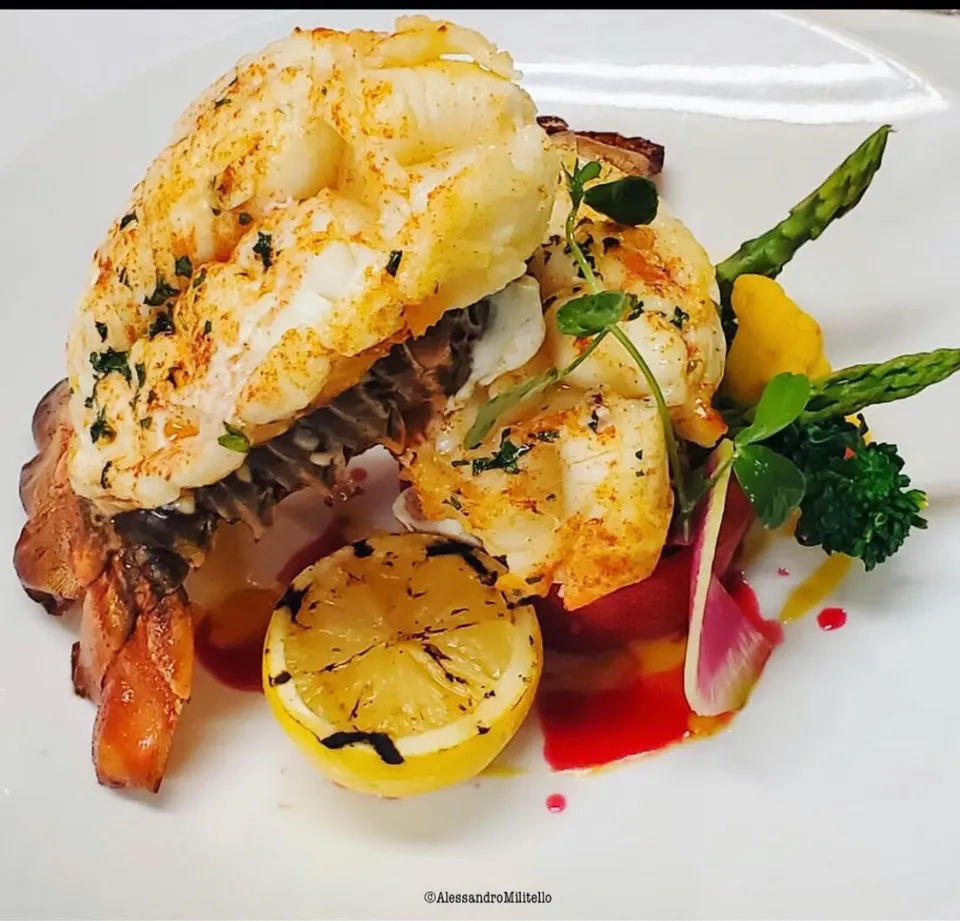 A beautifully plated lobster tail dish served with grilled lemon, asparagus spears, and vibrant garnishes on a white plate.