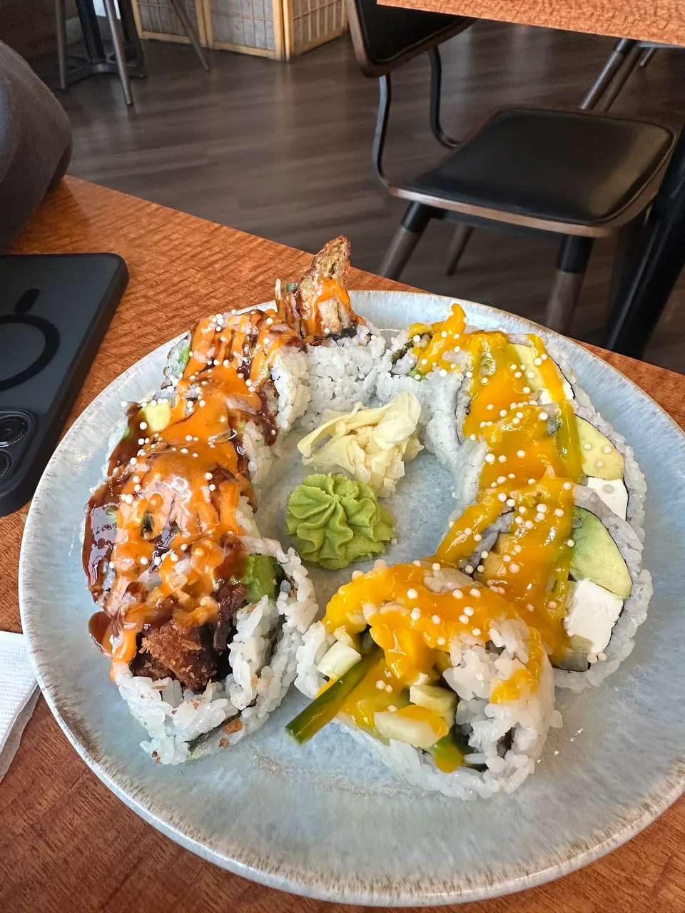 A plate of sushi rolls arranged in a circle, featuring one roll topped with a spicy orange sauce and another with a bright yellow mango sauce, served with ginger and wasabi in the center.