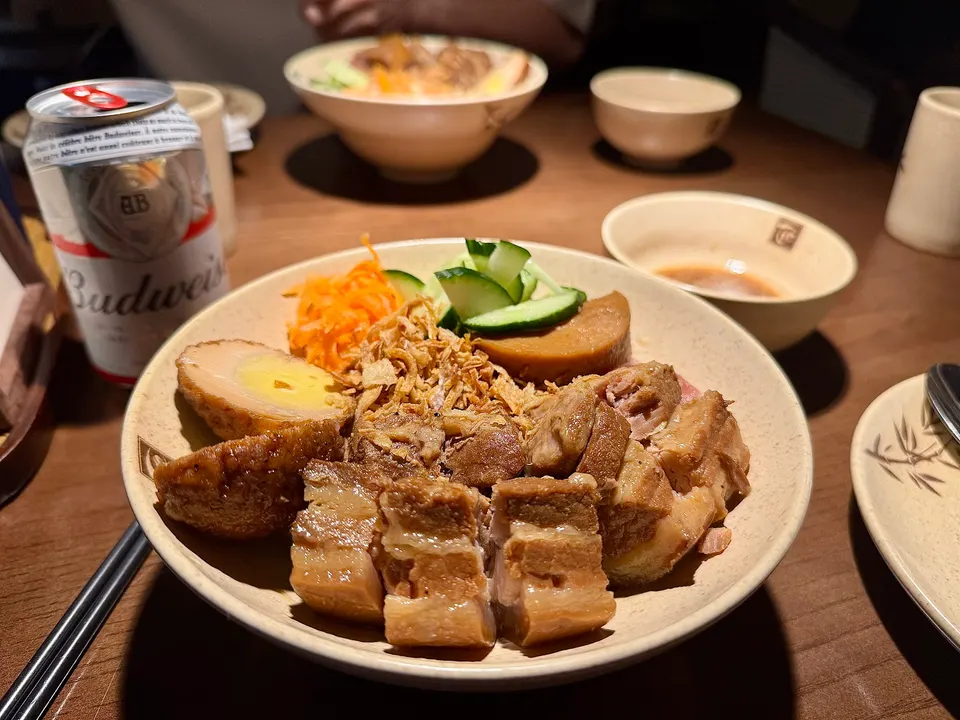 A bowl of Vietnamese braised pork belly with a soft-boiled egg, pickled carrots, cucumbers, and fried shallots, served with a can of Budweiser beer on a wooden table.