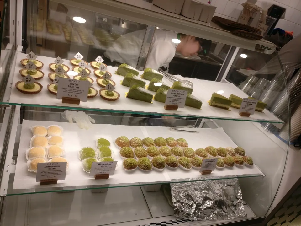 A glass display case at Tsujiri Dundas filled with Japanese matcha desserts, including small tarts, sliced green tea cakes, and rows of round mochi in various shades of green and beige.