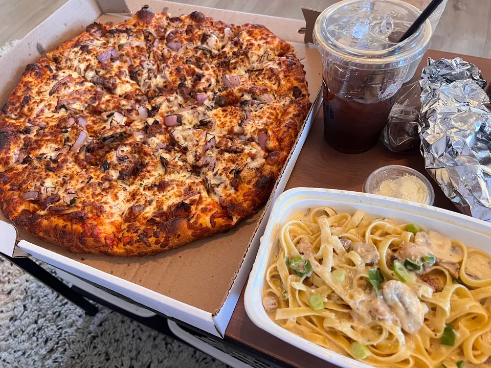 A large pizza with red onions and mushrooms in a cardboard box next to a container of creamy fettuccine pasta, an iced drink, and wrapped food.