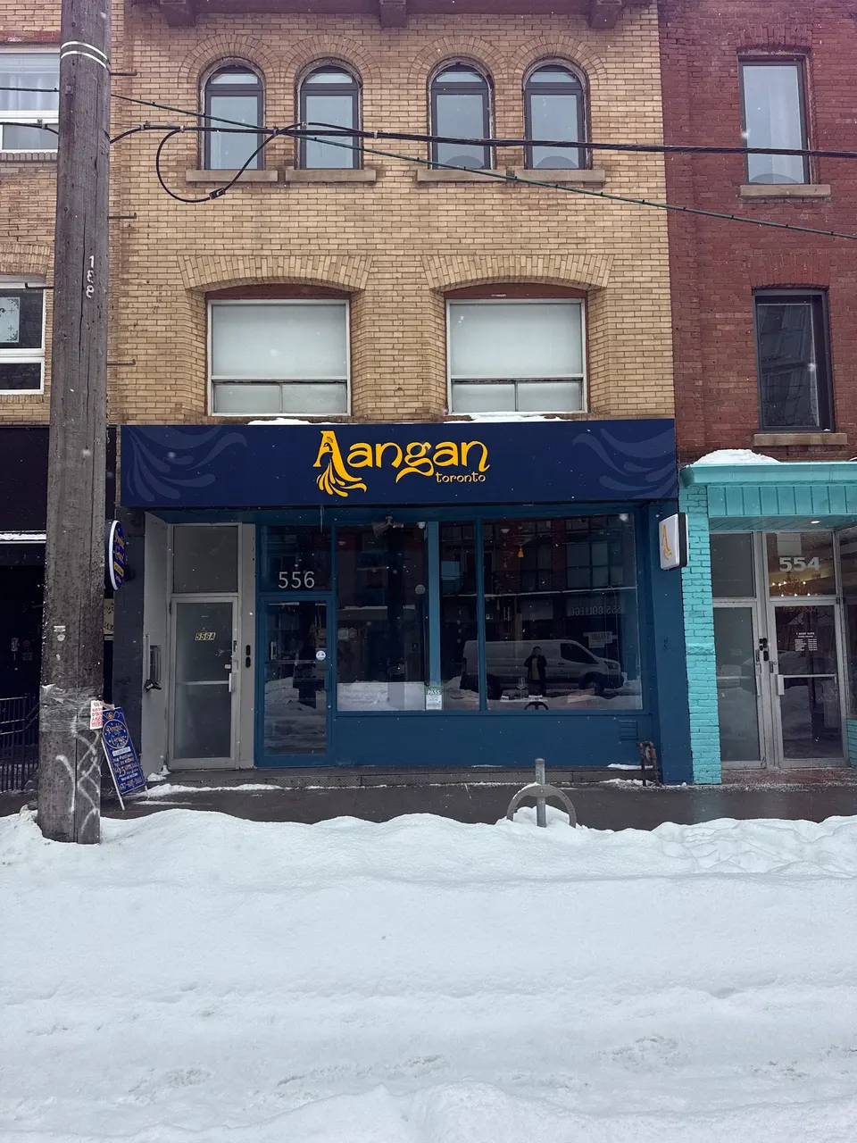 The exterior storefront of Aangan Toronto, an Indian restaurant, featuring a dark blue sign with yellow lettering, set against a brick building with snow piled on the sidewalk in front.