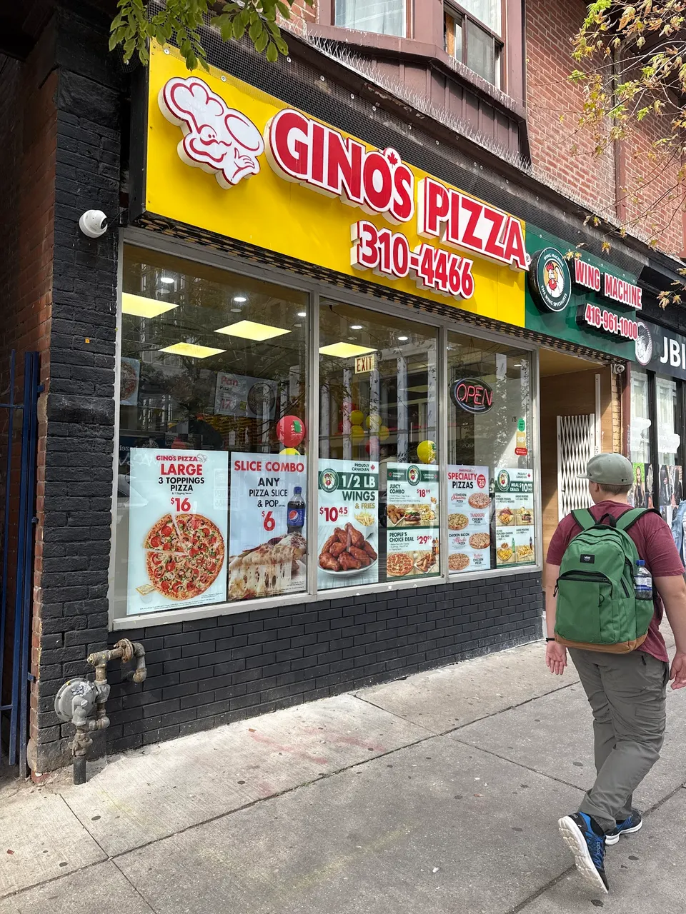 Exterior storefront of Gino's Pizza and Wing Machine on a city street, featuring a bright yellow sign with red lettering, window posters showing pizza and wing specials, and a pedestrian walking past.