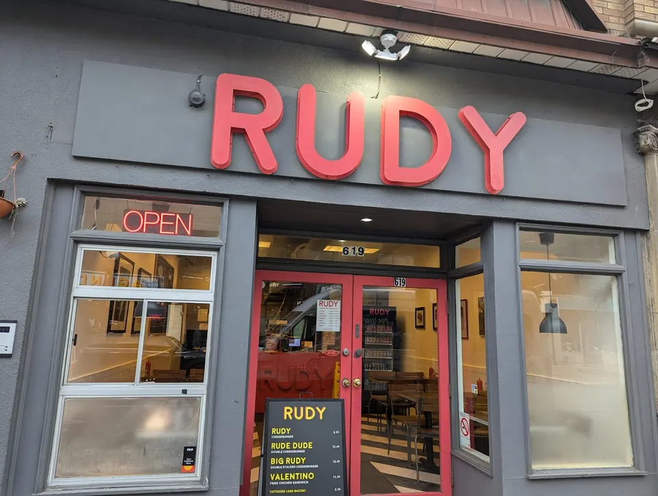 Exterior storefront of RUDY restaurant with a large red sign, glass windows, a red double door entrance, and a black sidewalk menu board displaying burger options.
