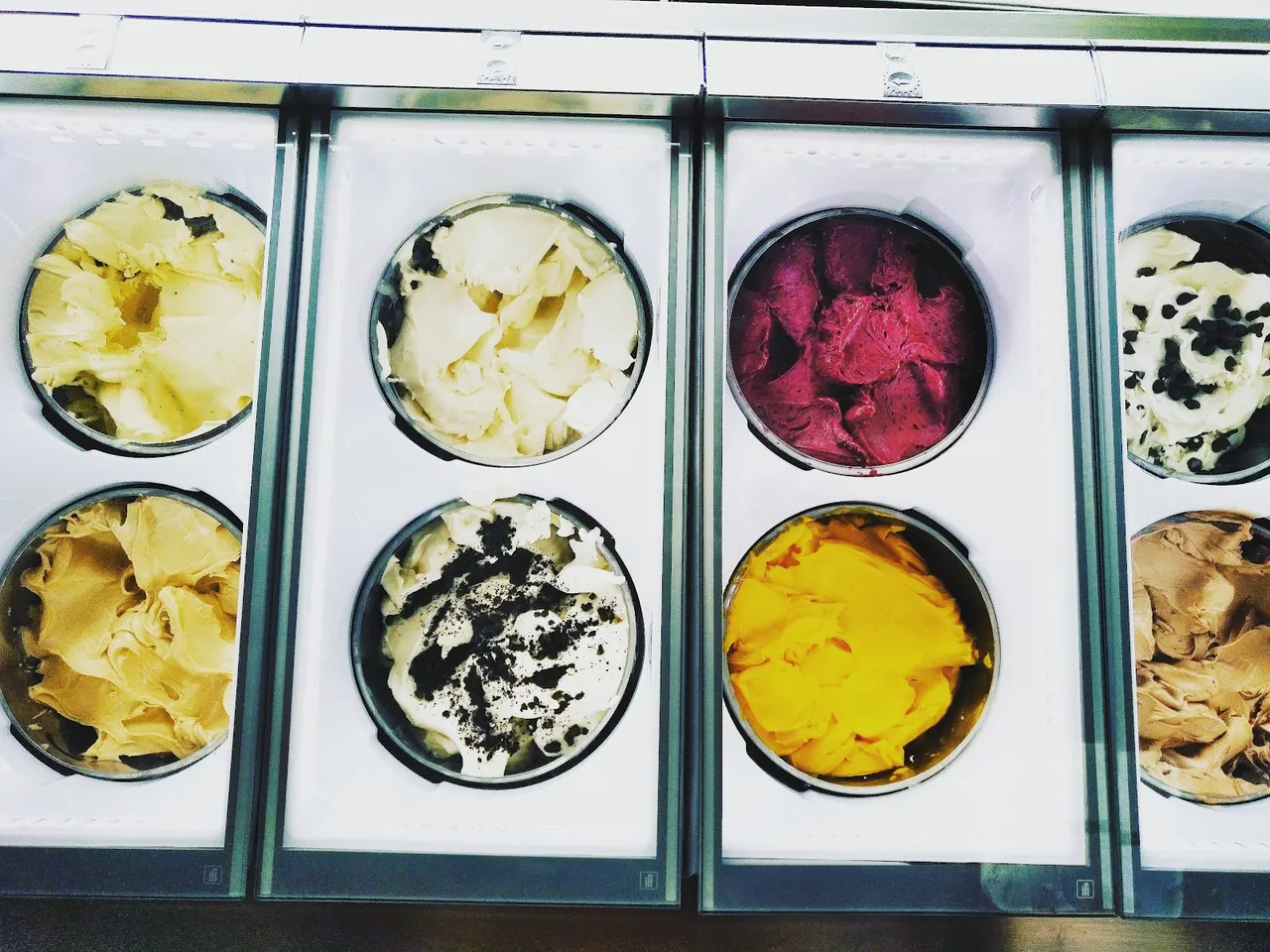 Eight tubs of colorful gelato displayed in a clean, modern freezer case, featuring flavors like mango, berry, chocolate chip, and cookies and cream.