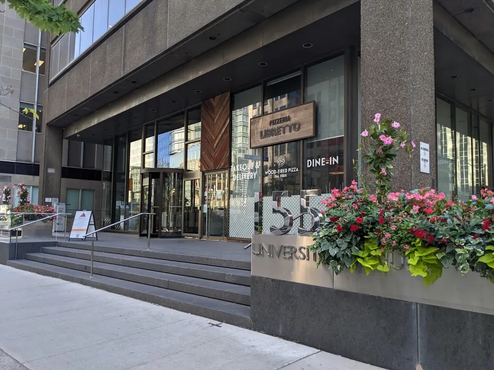 Exterior view of Pizzeria Libretto on University Avenue, featuring a modern concrete facade, steps leading to the entrance, a wooden sign, and a large planter with pink flowers.