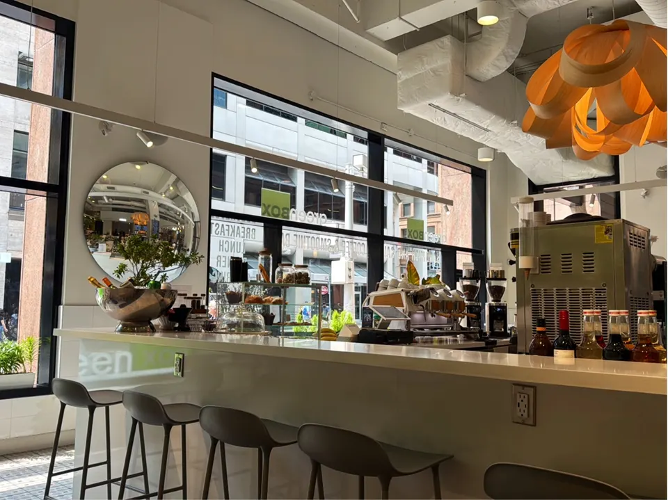 A modern cafe counter at Green Box with sleek bar stools, a white countertop, coffee machines, and large windows looking out onto a city street.