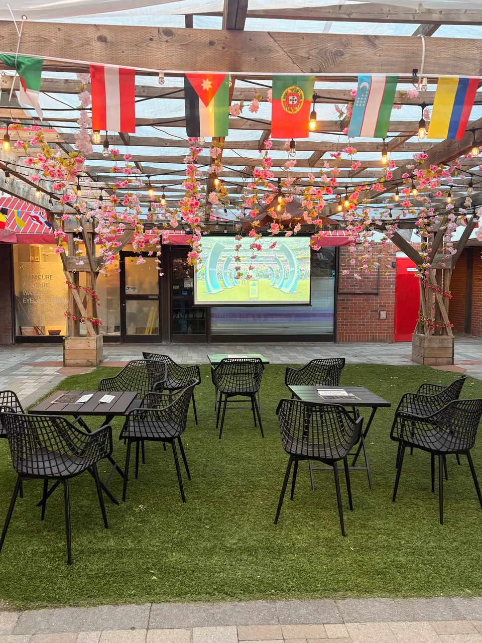An outdoor patio dining area at Capybara with artificial grass, black wire chairs, tables, a projector screen showing a game, and pink floral decorations hanging from a wooden pergola.