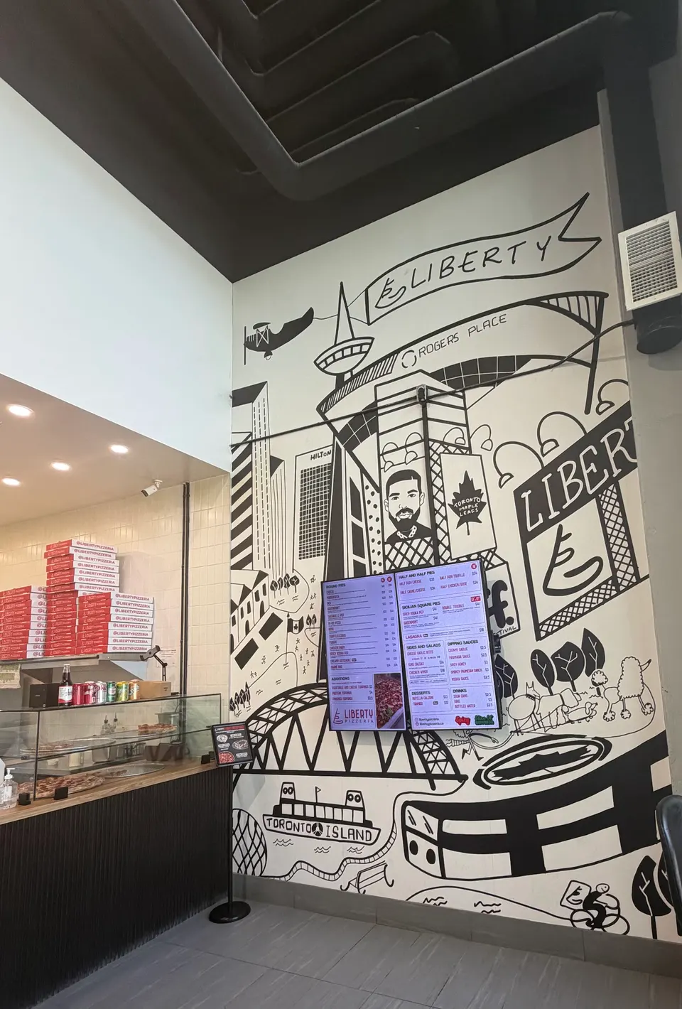 Interior of Liberty Pizzeria showing a large black and white wall mural depicting Toronto landmarks, digital menu screens, and a pizza counter with stacked boxes.