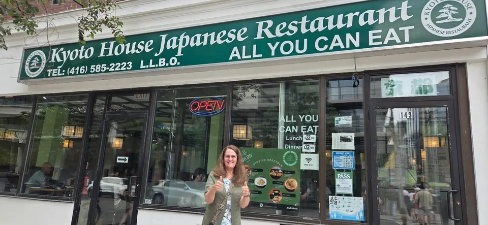 A woman stands smiling with thumbs up in front of Kyoto House Japanese Restaurant, featuring a green storefront sign advertising all-you-can-eat sushi.
