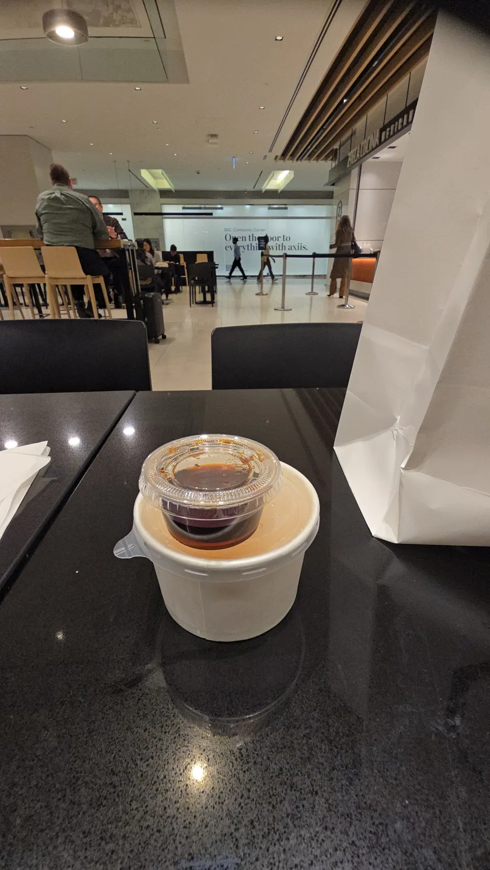 A takeout container of soup topped with a small plastic cup of dark dipping sauce, sitting on a black table at Zen Kyoto in a food court setting.