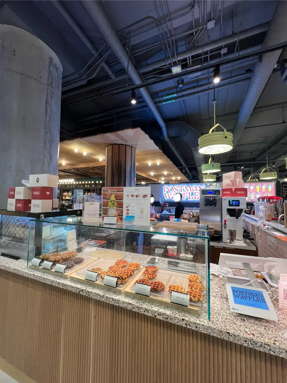 Interior view of Postman Waffles cafe, showing a display case with various waffles, a counter with waffle-making equipment, and a neon sign in the background. The ceiling has exposed pipes and light fixtures.