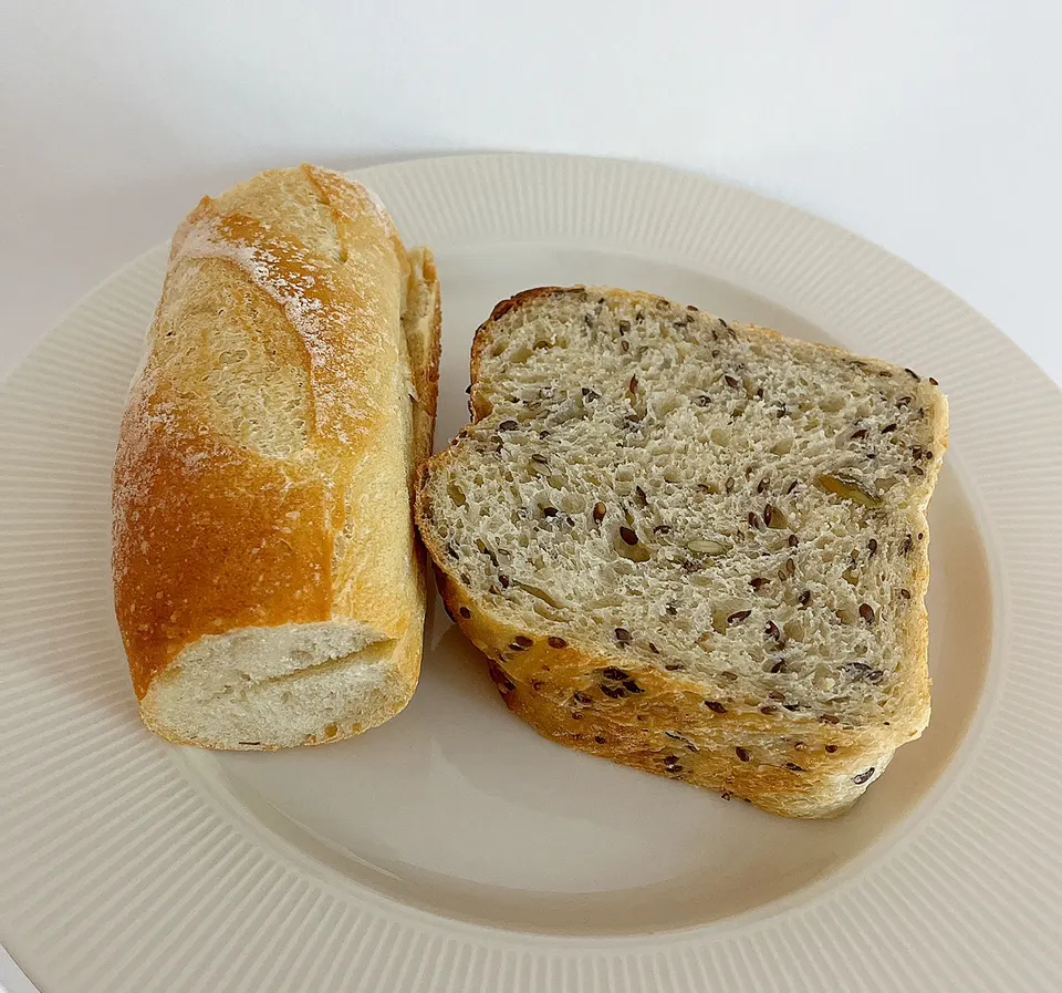 A slice of seeded multigrain bread and a small crusty baguette roll served side-by-side on a white ribbed ceramic plate.
