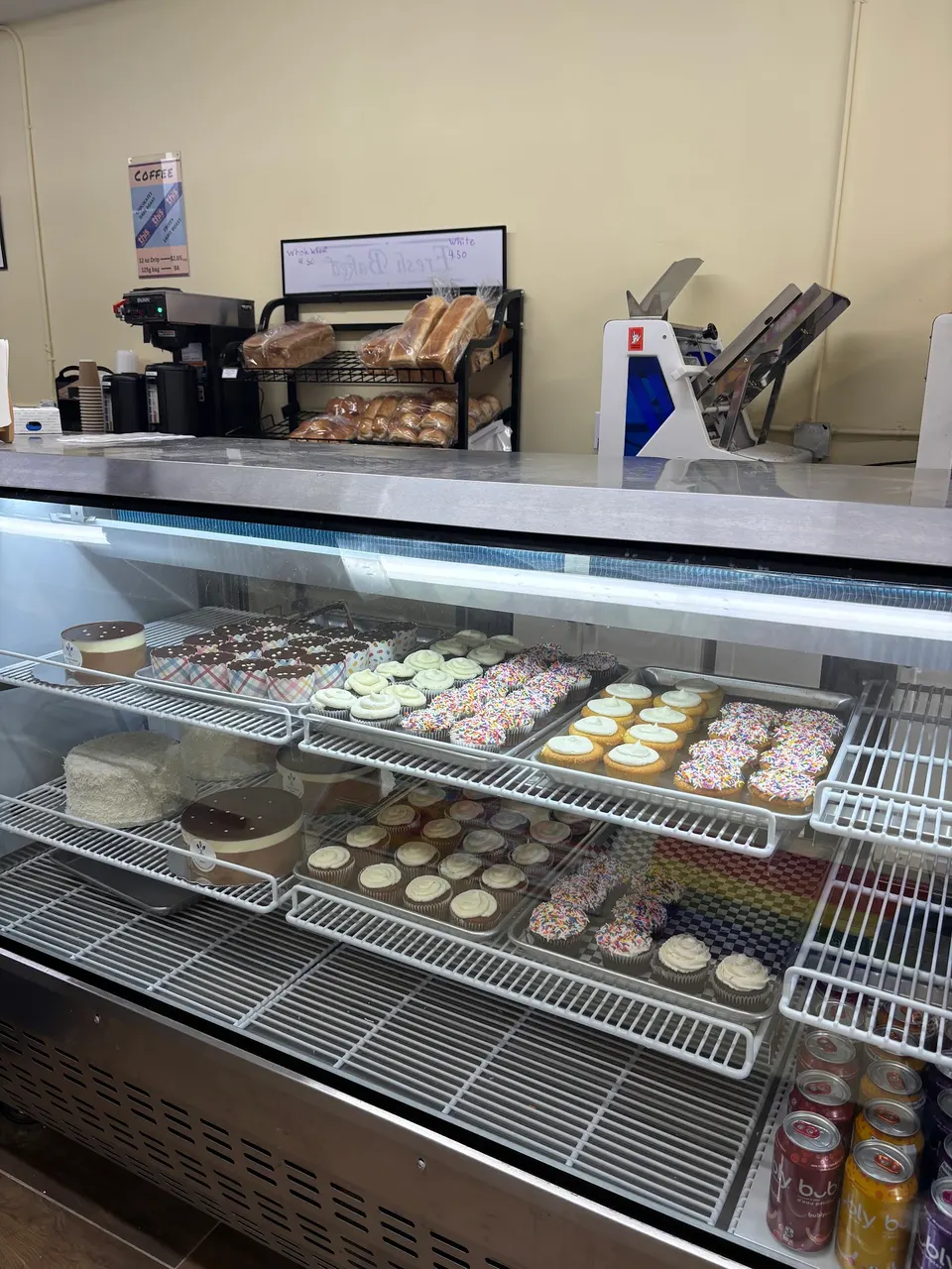 A refrigerated bakery display case filled with various cupcakes, cakes, and pastries, with bread loaves on a shelf and coffee equipment in the background.