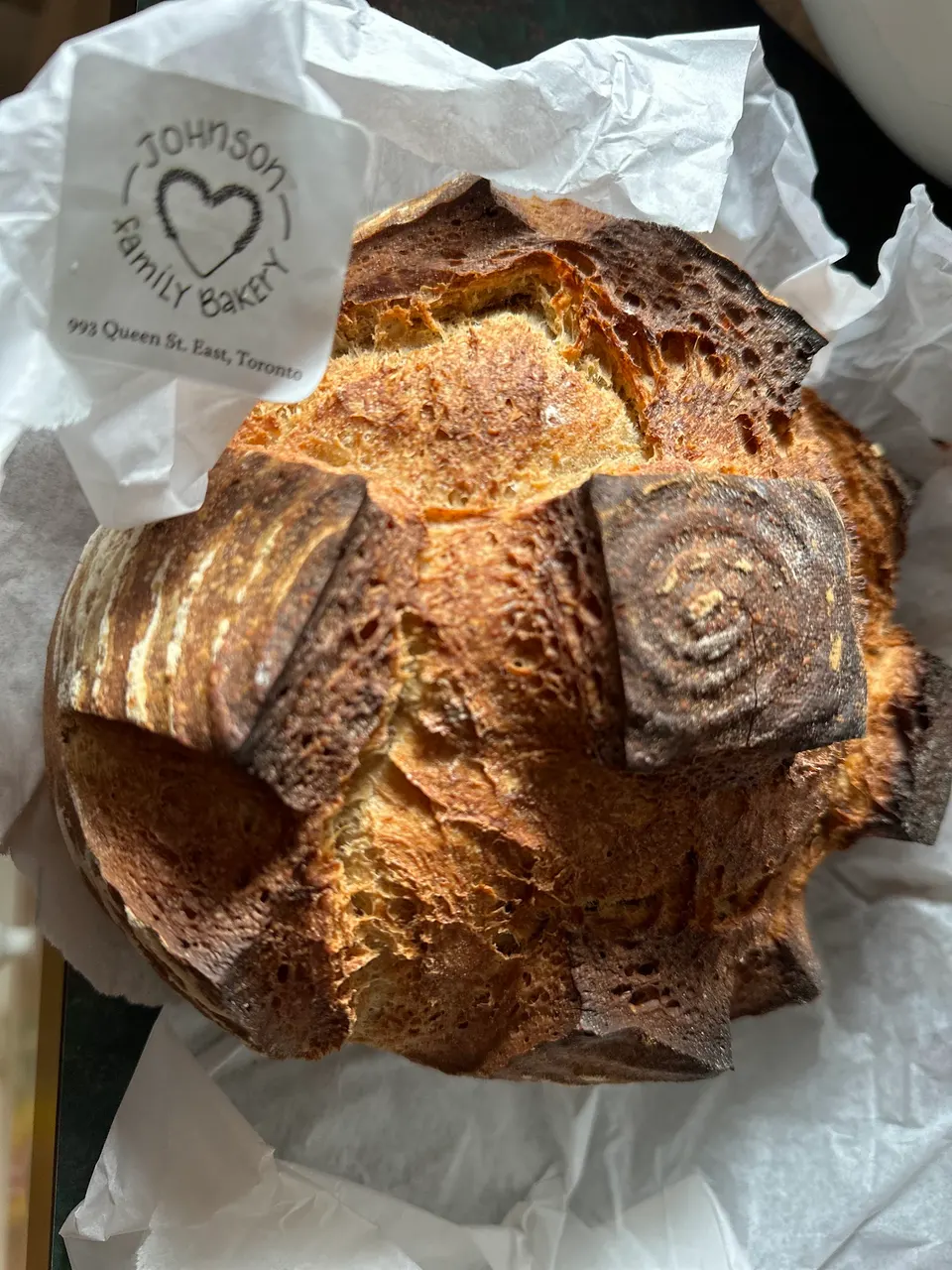 A freshly baked, crusty sourdough bread loaf with a cross-cut pattern on top, resting in white parchment paper with a Johnson Family Bakery sticker visible.