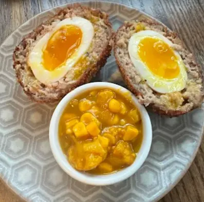 A soft-boiled Scotch egg cut in half, showcasing a runny golden yolk, served on a patterned plate alongside a small white bowl of yellow piccalilli relish.