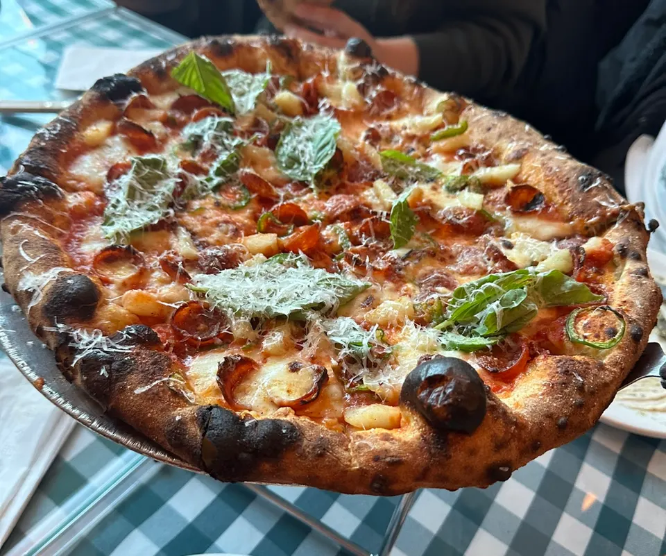 A freshly baked wood-fired pizza on a metal stand, topped with pepperoni, pineapple chunks, fresh basil leaves, and grated parmesan cheese.