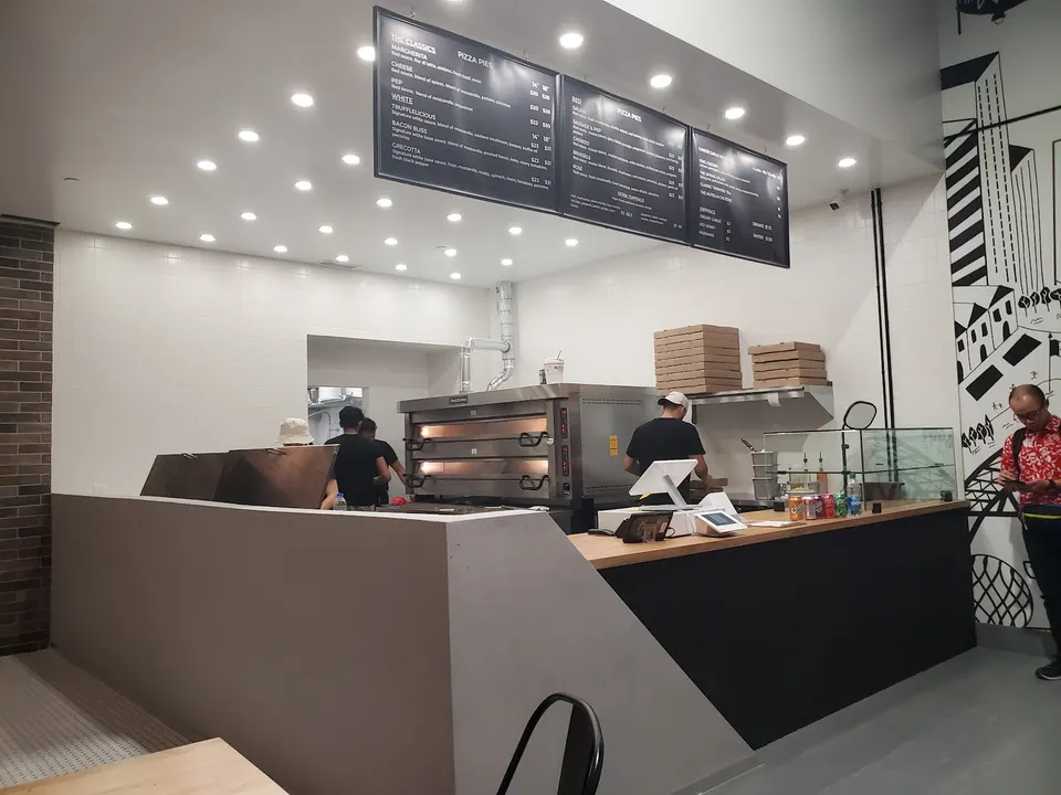 Interior view of Liberty Pizzeria showing the service counter, pizza ovens, staff preparing orders, overhead menu boards, and a customer waiting on the right.