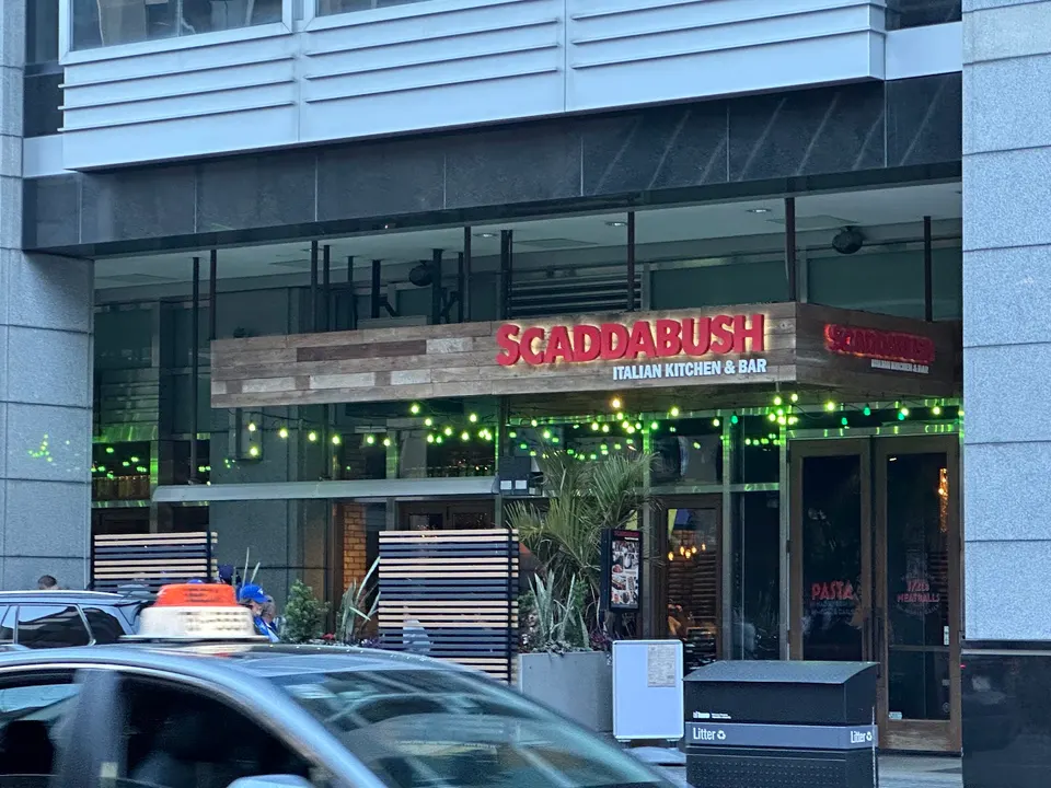 The exterior entrance of Scaddabush Italian Kitchen & Bar on Front Street, featuring a wooden sign with red lettering, green string lights, and patio dividers.