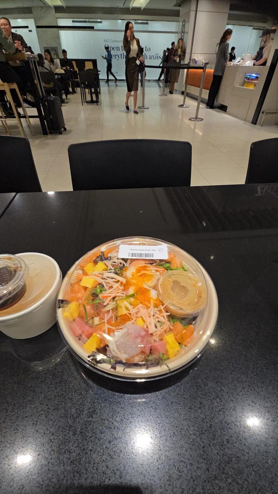 A packaged sashimi salad bowl and a side container on a dark counter at Zen Kyoto, with a blurred background showing people walking in a modern food court setting.