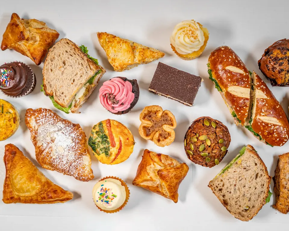 An overhead view of an assortment of baked goods from Mabel's Bakery, including sandwiches, croissants, muffins, cupcakes, and savory pastries arranged on a white surface.