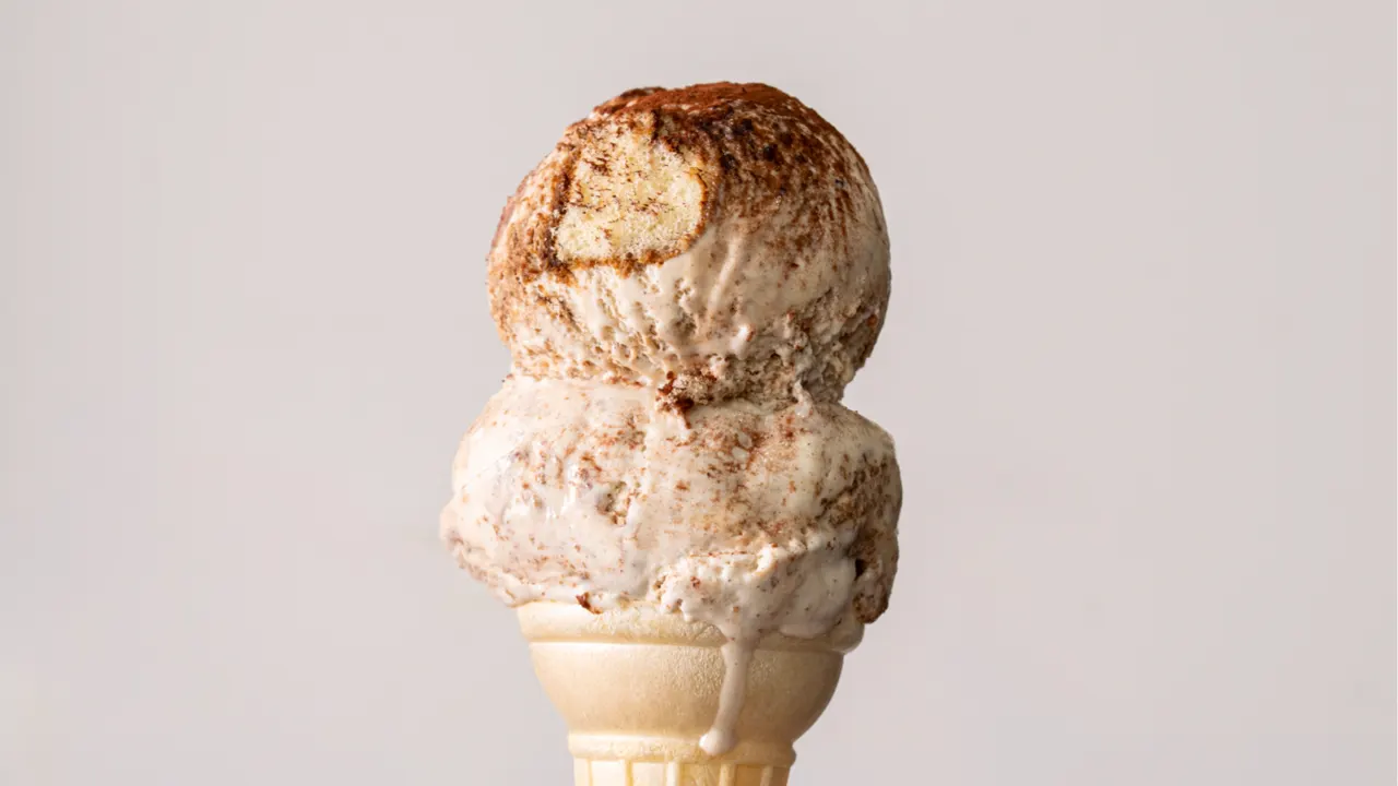 Two scoops of tiramisu ice cream dusted with cocoa powder on a wafer cone against a plain light grey background.