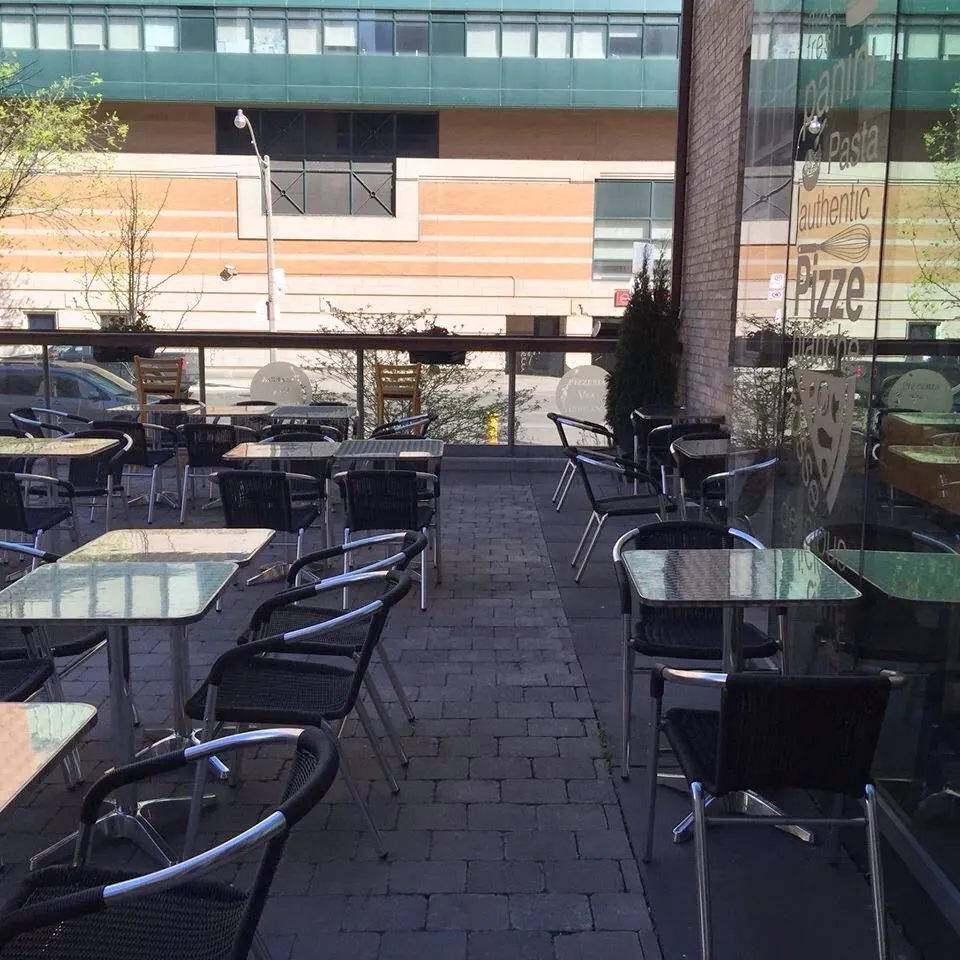 An outdoor patio dining area at Pizzeria Via Mercanti with metal and black wicker chairs, square glass-topped tables, and a brick-paved floor next to a glass window with restaurant branding.