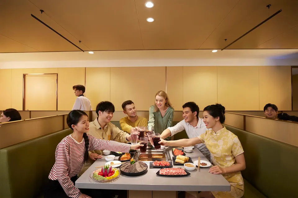 A group of friends smiling and clinking glasses together over a hot pot table filled with fresh ingredients at Haidilao Hot Pot Toronto Downtown.