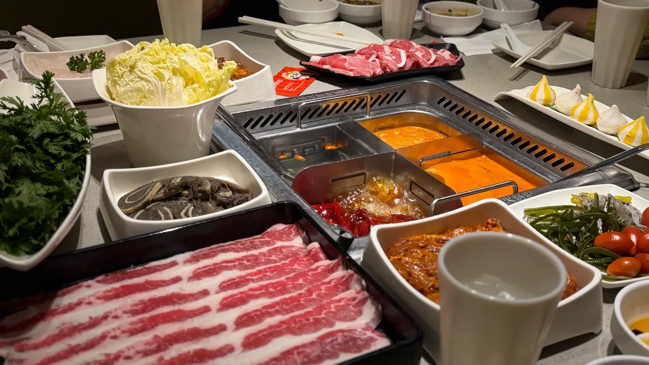 A table spread at Haidilao Hot Pot featuring a multi-compartment hot pot with bubbling broths, surrounded by raw sliced beef, leafy greens, napa cabbage, and various side dishes.
