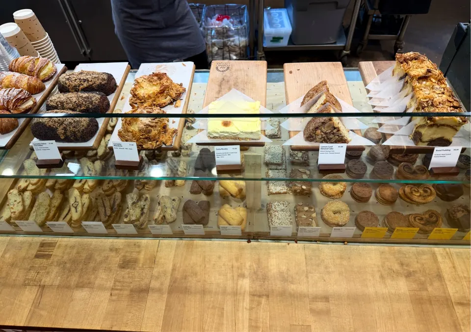 A glass display case at Forno Cultura bakery filled with various Italian pastries, including croissants, biscotti, cakes, and cookies on wooden boards.