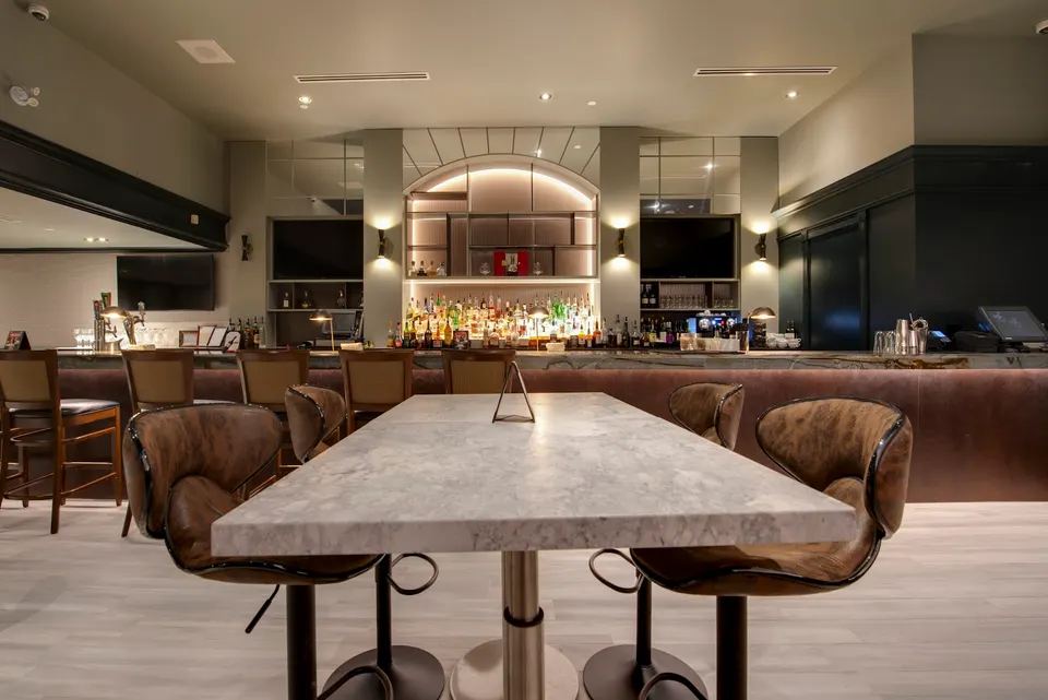 A modern bar area with a marble-topped high-top table in the foreground, surrounded by brown leather barstools, looking toward a well-lit bar counter with liquor bottles and wall-mounted TVs.