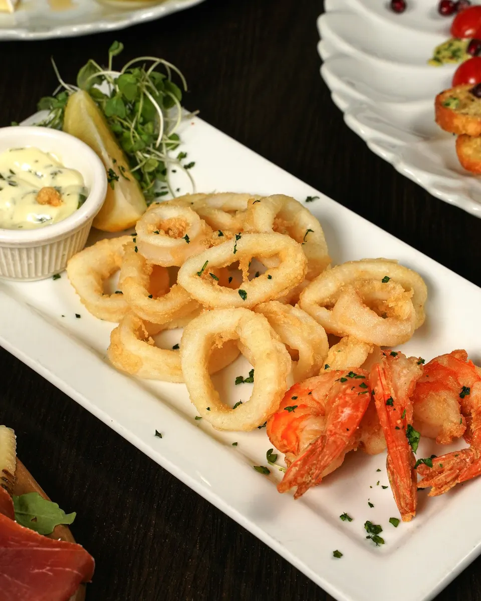 A long white platter presenting crispy golden-brown fried calamari rings and fried shrimp, served with a side of creamy dipping sauce, a lemon wedge, and fresh microgreens.