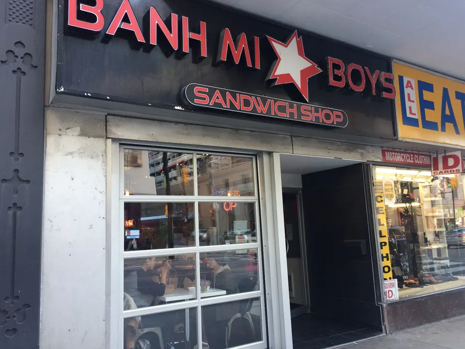The exterior storefront of Banh Mi Boys sandwich shop, featuring a large black sign with red and white lettering and a star logo above a glass garage-door style window.