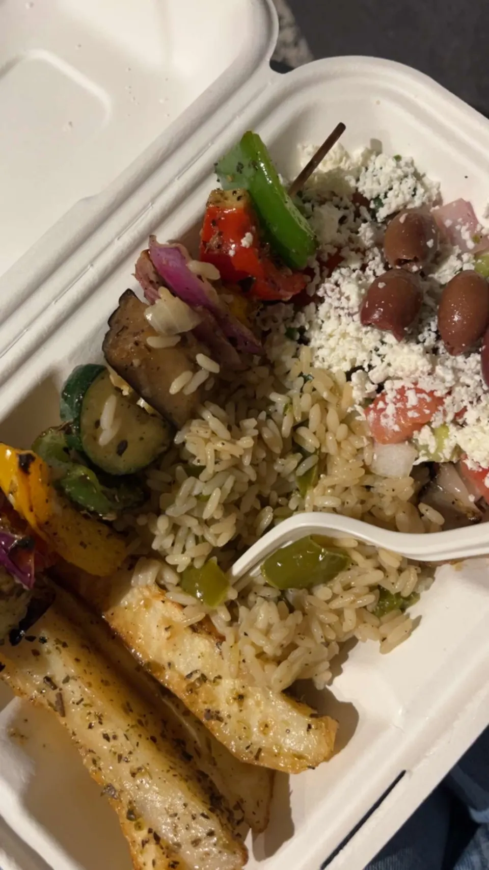 A Greek takeout container filled with a grilled vegetable skewer, seasoned rice, roasted potato wedges, and a Greek salad topped with crumbled feta cheese and kalamata olives.