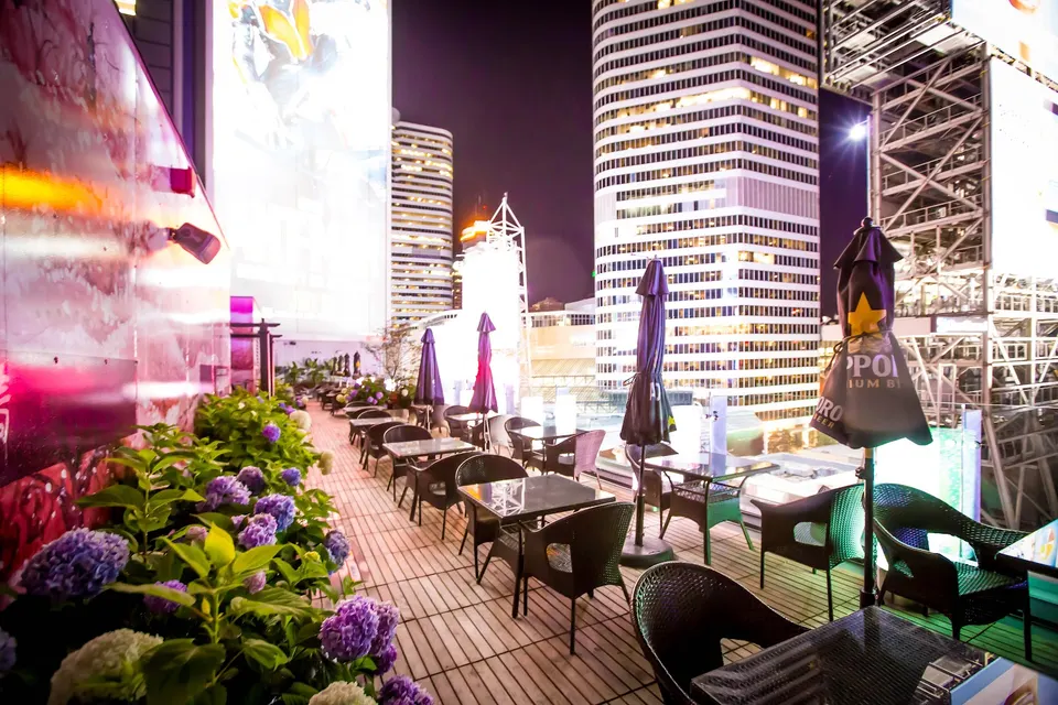 A rooftop patio at night with wooden decking, black dining tables and chairs, purple umbrellas, and lush planters of purple hydrangeas, set against a backdrop of illuminated skyscrapers.